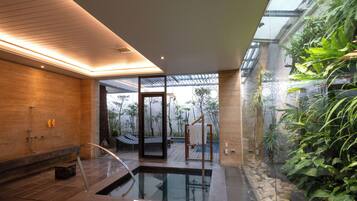 A modern spa area with a glass-enclosed hot tub, wooden flooring, and a view of a tropical garden.