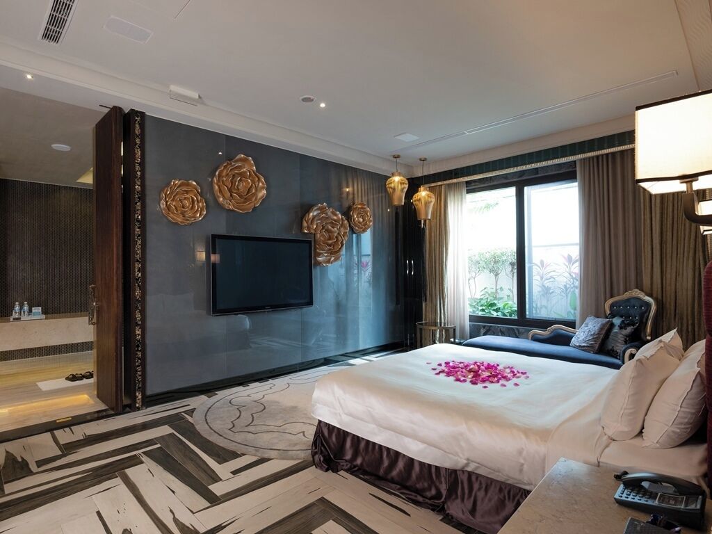 A hotel room with a large bed, a flat-screen TV, decorative wall art, and a view of greenery outside.