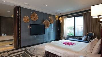 A hotel room with a large bed, a flat-screen TV, decorative wall art, and a view of greenery outside.