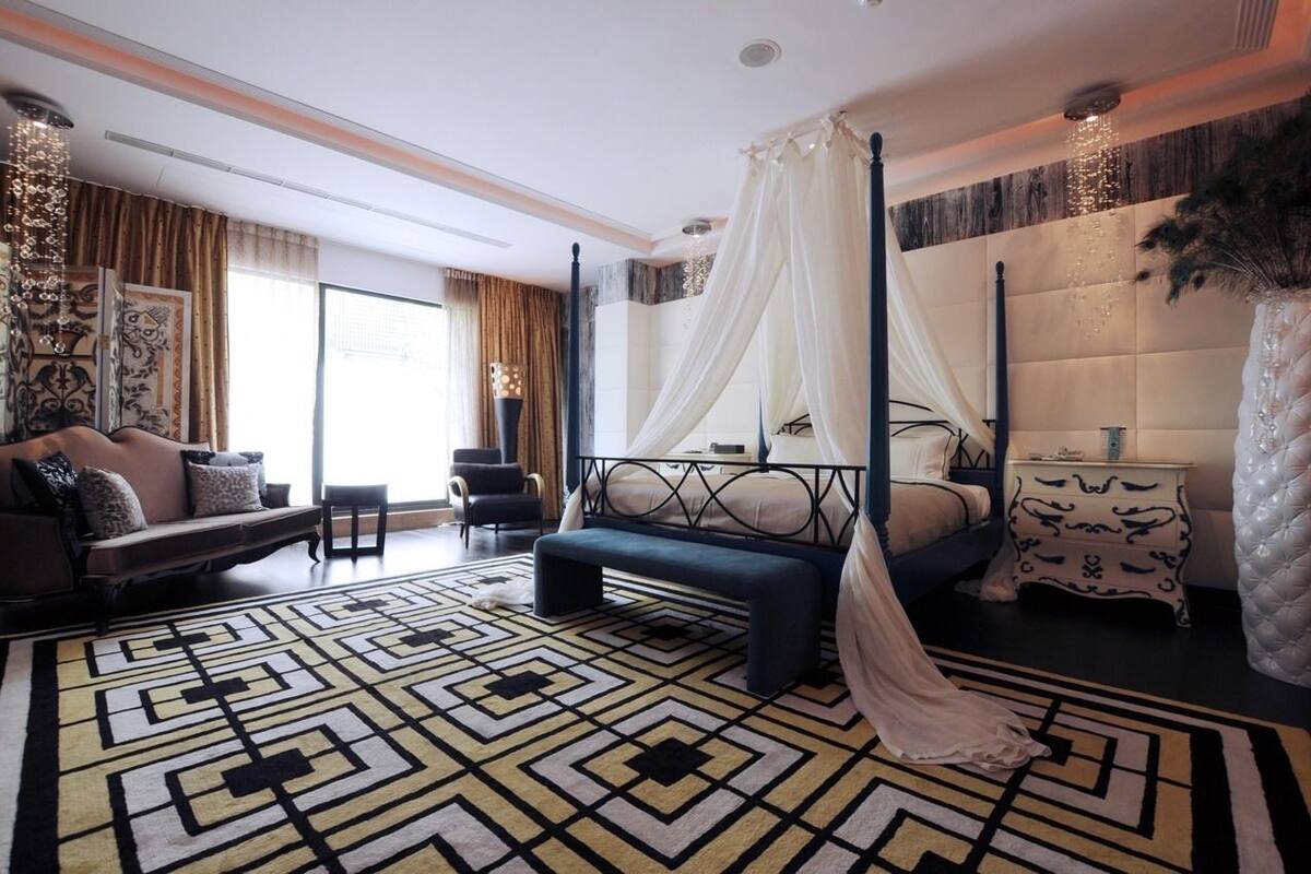 A bedroom with a four-poster bed, a sitting area with a sofa and armchairs, a patterned rug, and a decorative vase.