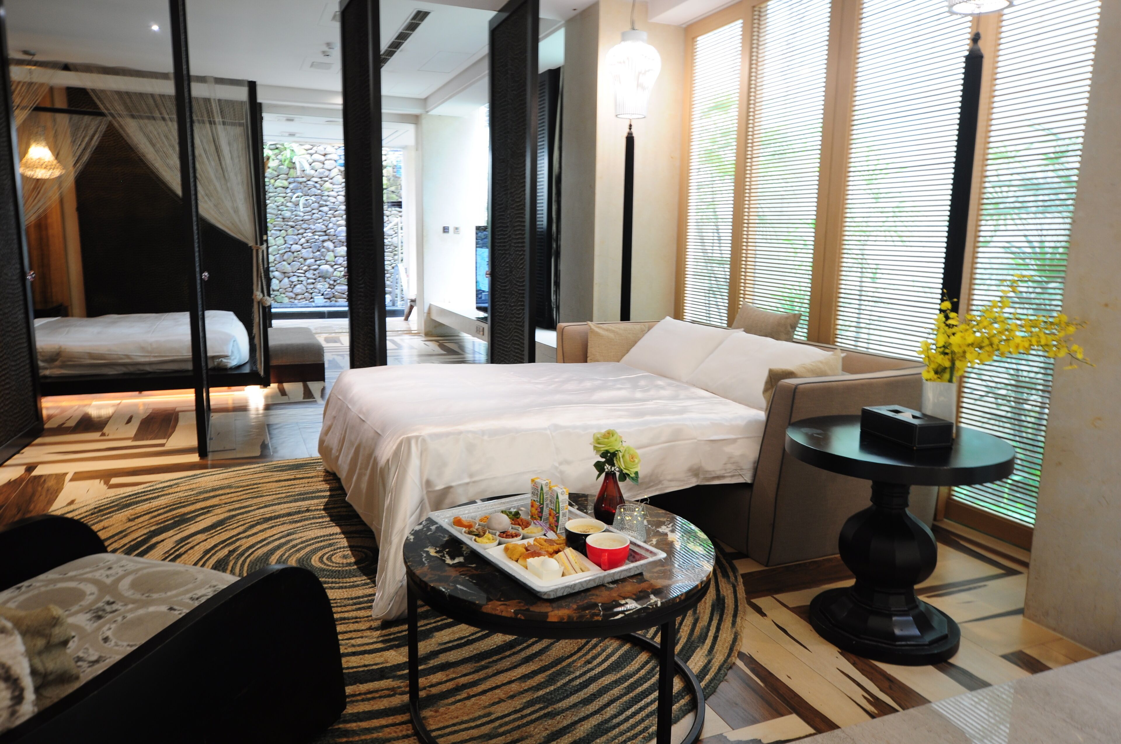 A hotel room with a bed, a sofa, a dining table with food, and a vase with flowers.