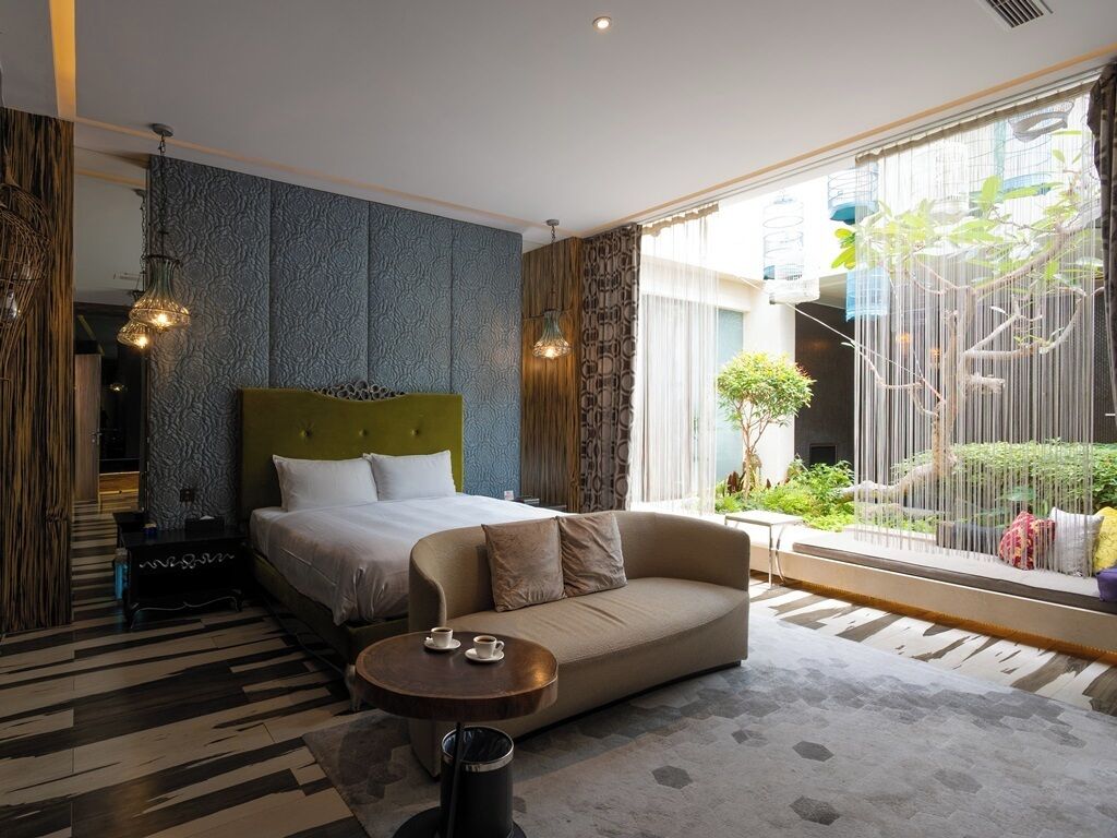 A modern hotel room with a large bed, a sofa, a coffee table, and a view of greenery outside.