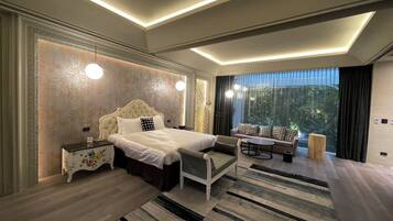 A modern bedroom with a large bed, bedside tables, a sofa, and a view of greenery through the window.