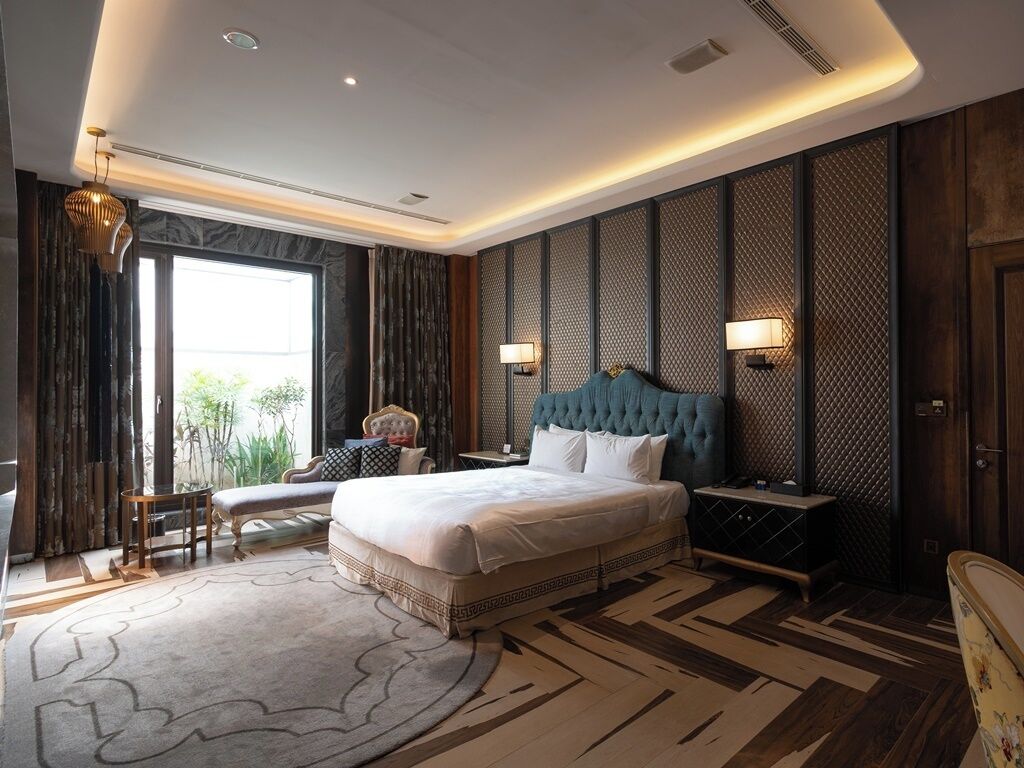A modern hotel room with a large bed, a desk, and a chair. There is a view of greenery outside the window.