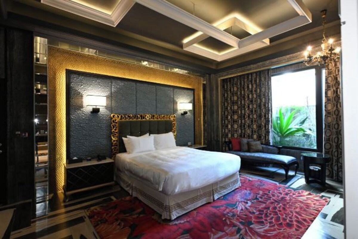 Habitación de hotel con una cama grande, un sofá, un tapete rojo estampado y vistas a la vegetación a través de la ventana.