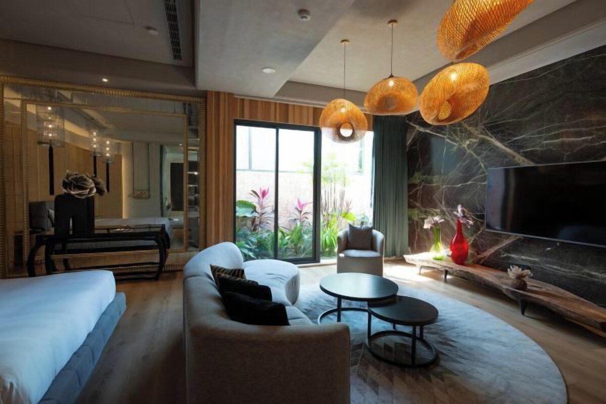 Una habitación de hotel moderna con una cama grande, un sofá, una mesa de comedor y un televisor. Cuenta con una chimenea y un ventanal con vistas a plantas.
