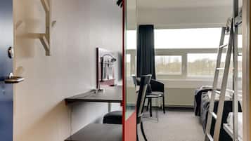 Quarto de hotel com escrivaninha, cadeira, cama e janela com vista para árvores.
