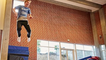 Una persona saltando en un gimnasio con paredes de ladrillo, ventanas grandes y colchonetas acolchadas.