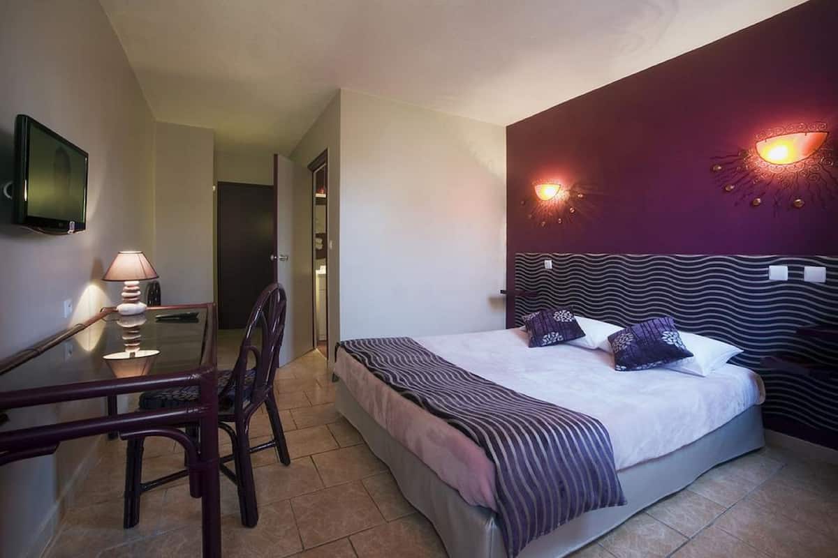 A hotel room with a bed, a desk with a lamp, and a TV mounted on the wall.