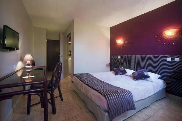 A hotel room with a bed, a desk with a lamp, and a TV mounted on the wall. - Tropic Hotel (Rivesaltes)