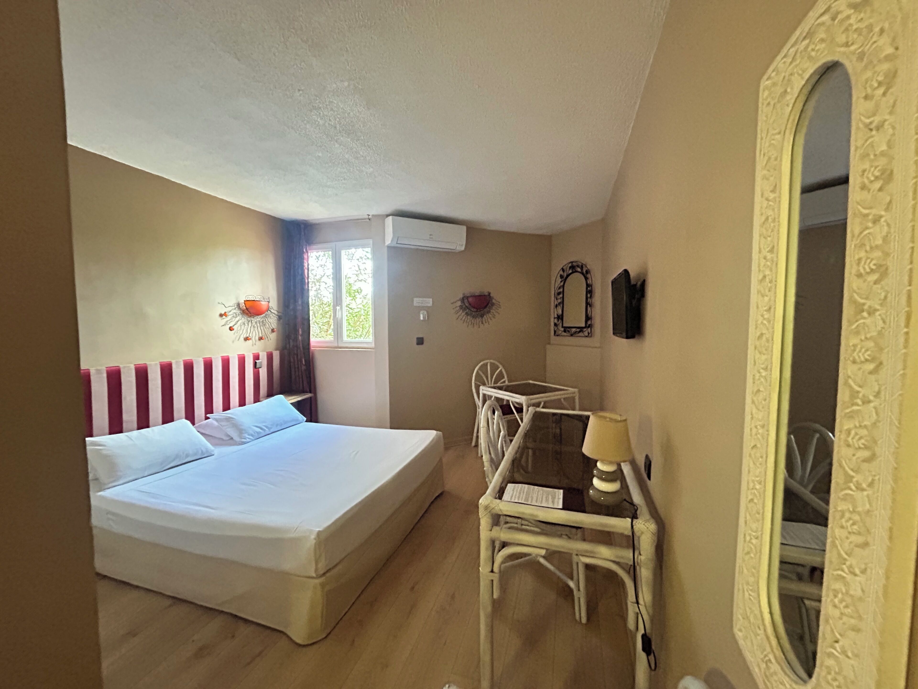 A bedroom with a bed, a small table, a chair, a mirror, and a window.