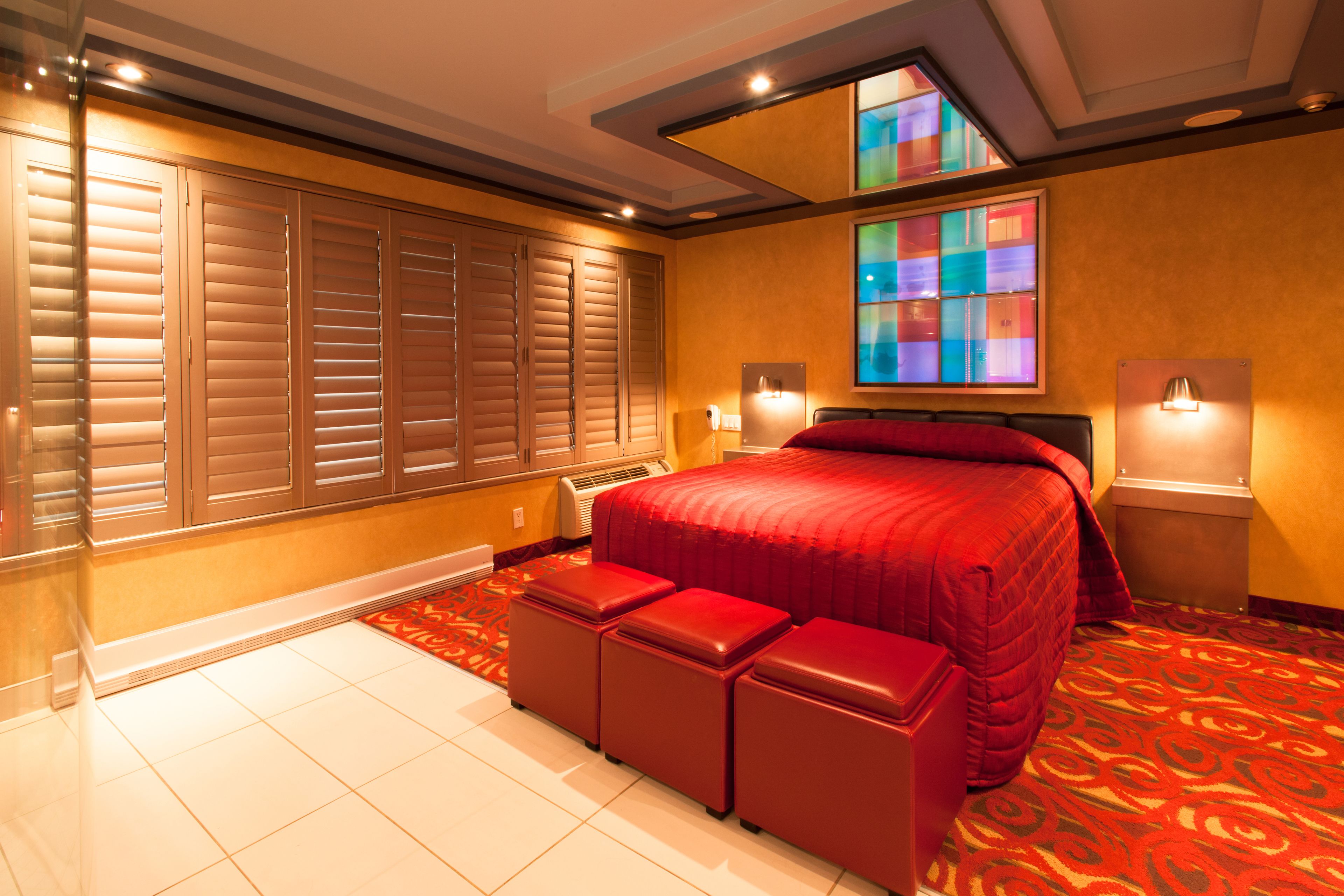 Una habitación de hotel con una cama grande, ropa de cama roja, un otomano rojo, un ventanal con un diseño de vitral y suelo de baldosas.