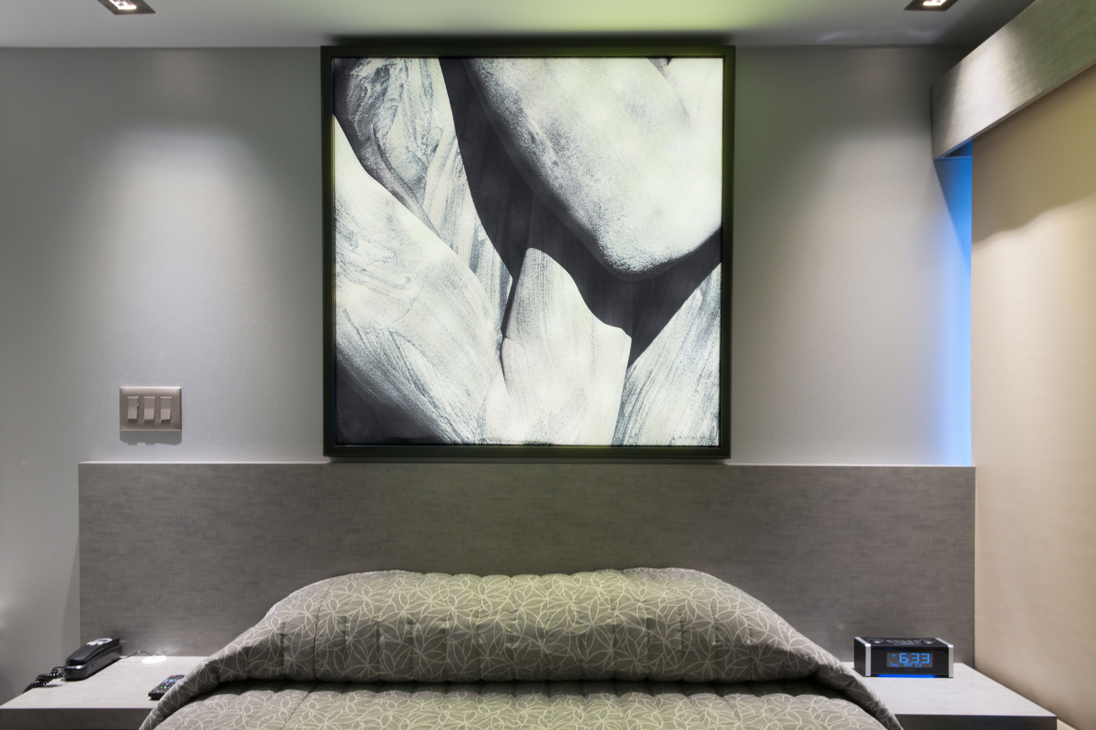 A modern hotel room with a large abstract painting on the wall, a neatly made bed with patterned bedding, a bedside table with a remote control, and a digital clock.