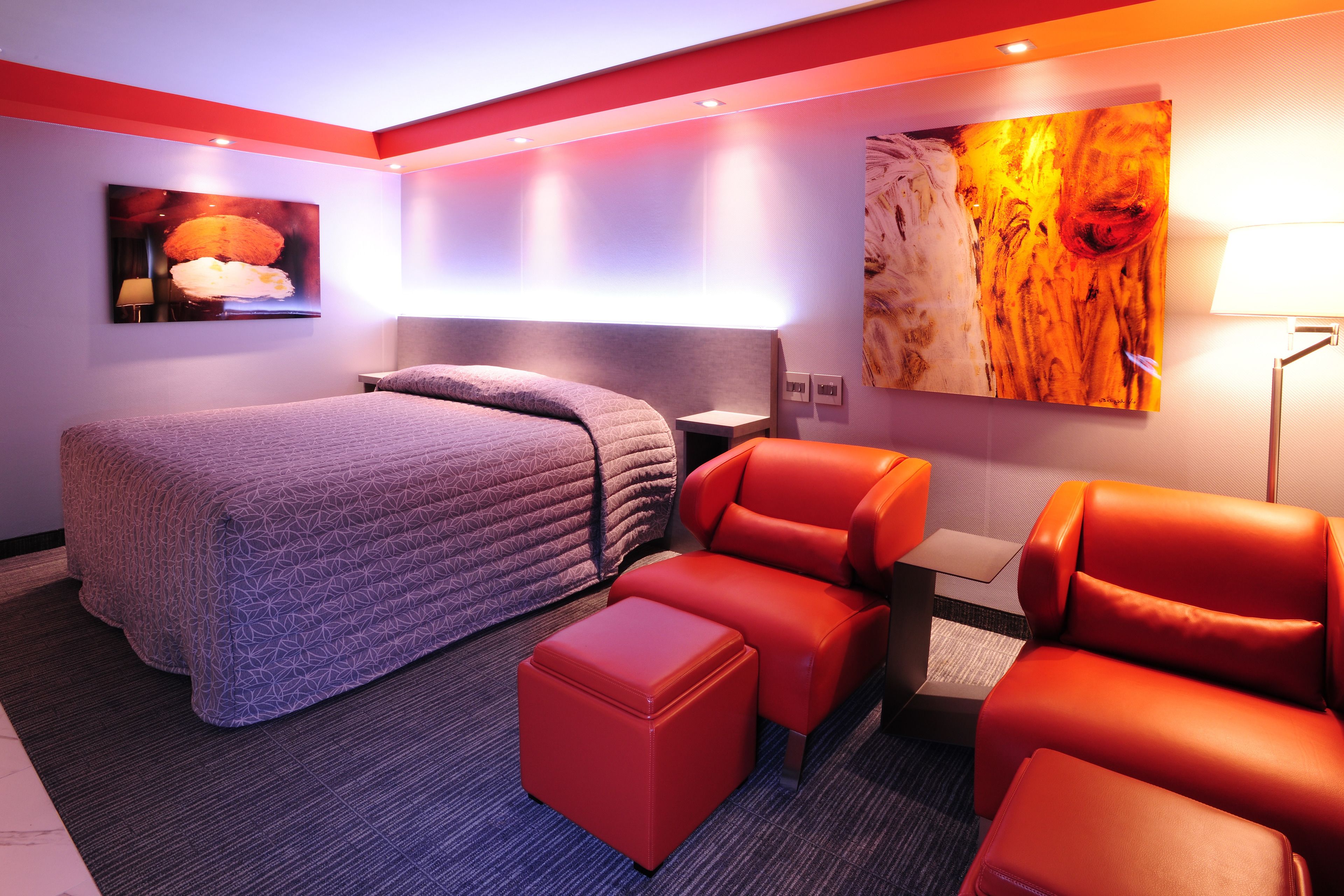 Ein modernes Hotelzimmer mit einem großen Bett, einem roten Ledersofa, einem kleinen Hocker, einem Nachttisch und einer Wandleuchte.