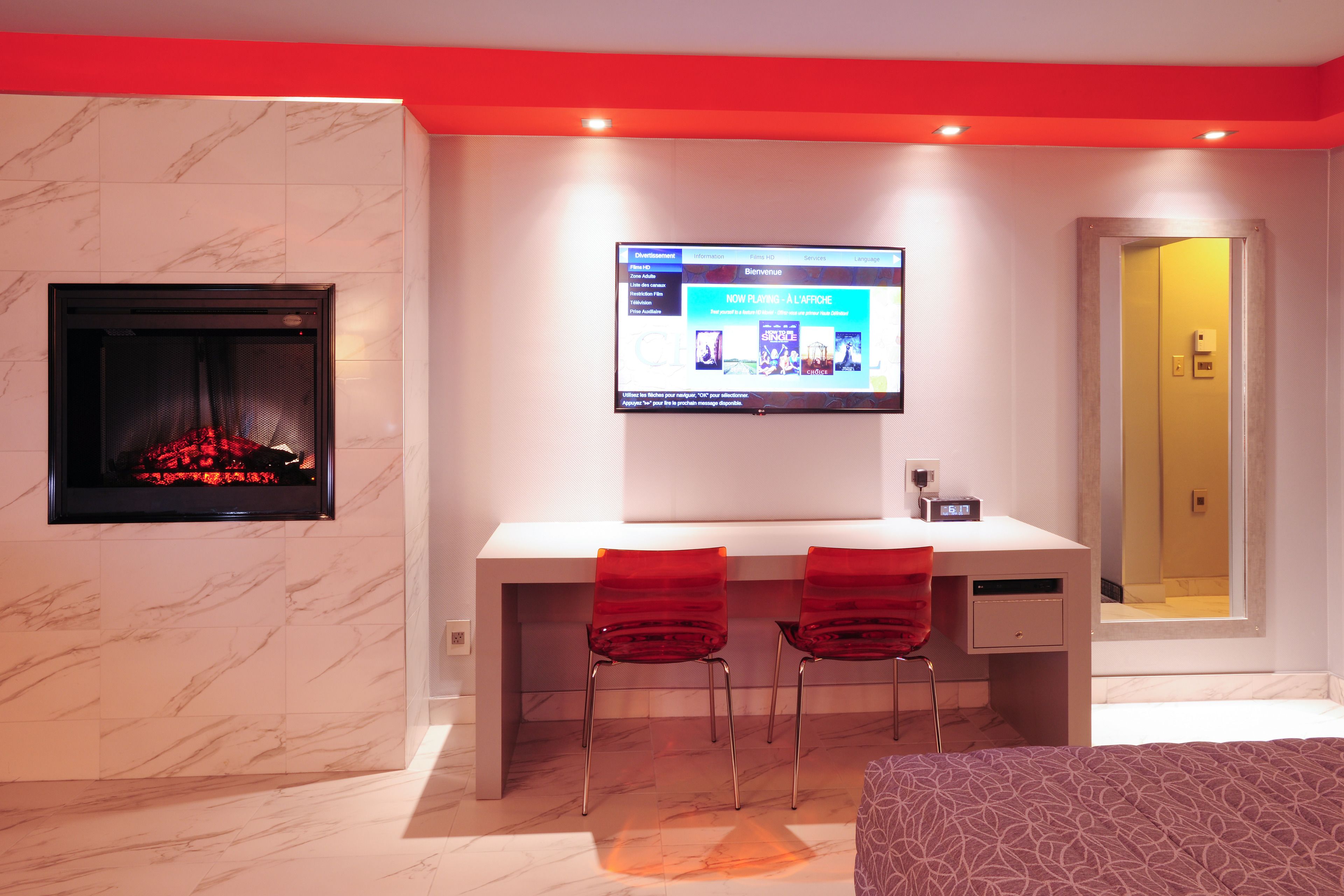 A modern hotel room with a fireplace, a desk with two red chairs, a television, and a mirror.