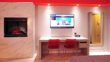 A modern hotel room with a fireplace, a desk with two red chairs, a television, and a mirror.