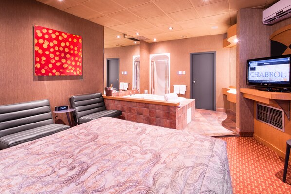 Habitación de hotel con una cama grande, zona de estar con televisor, baño con ducha y pared con estampado rojo y naranja.