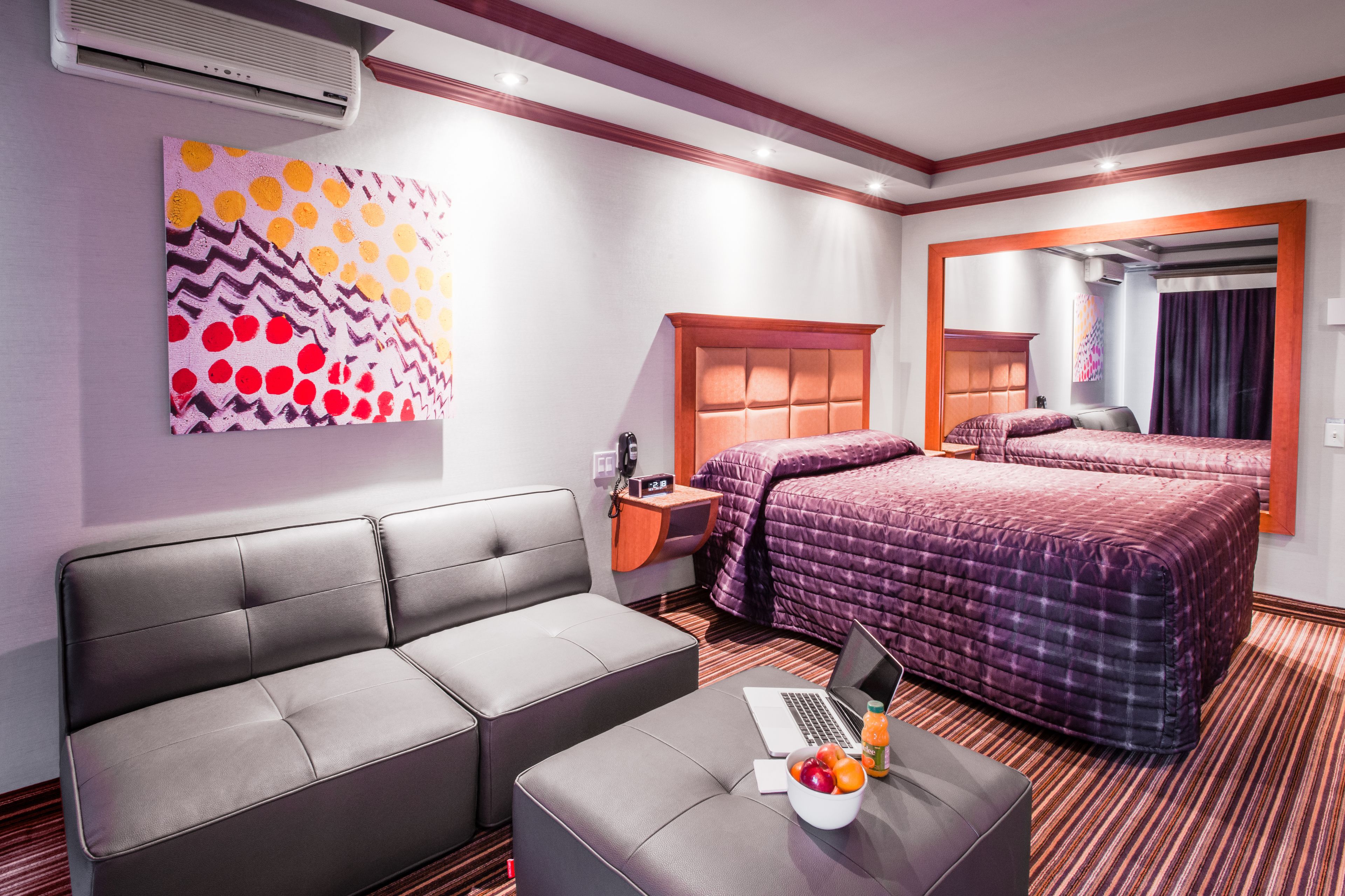 Habitación de hotel con sofá, cama con colcha morada, laptop sobre una mesa y frutero.