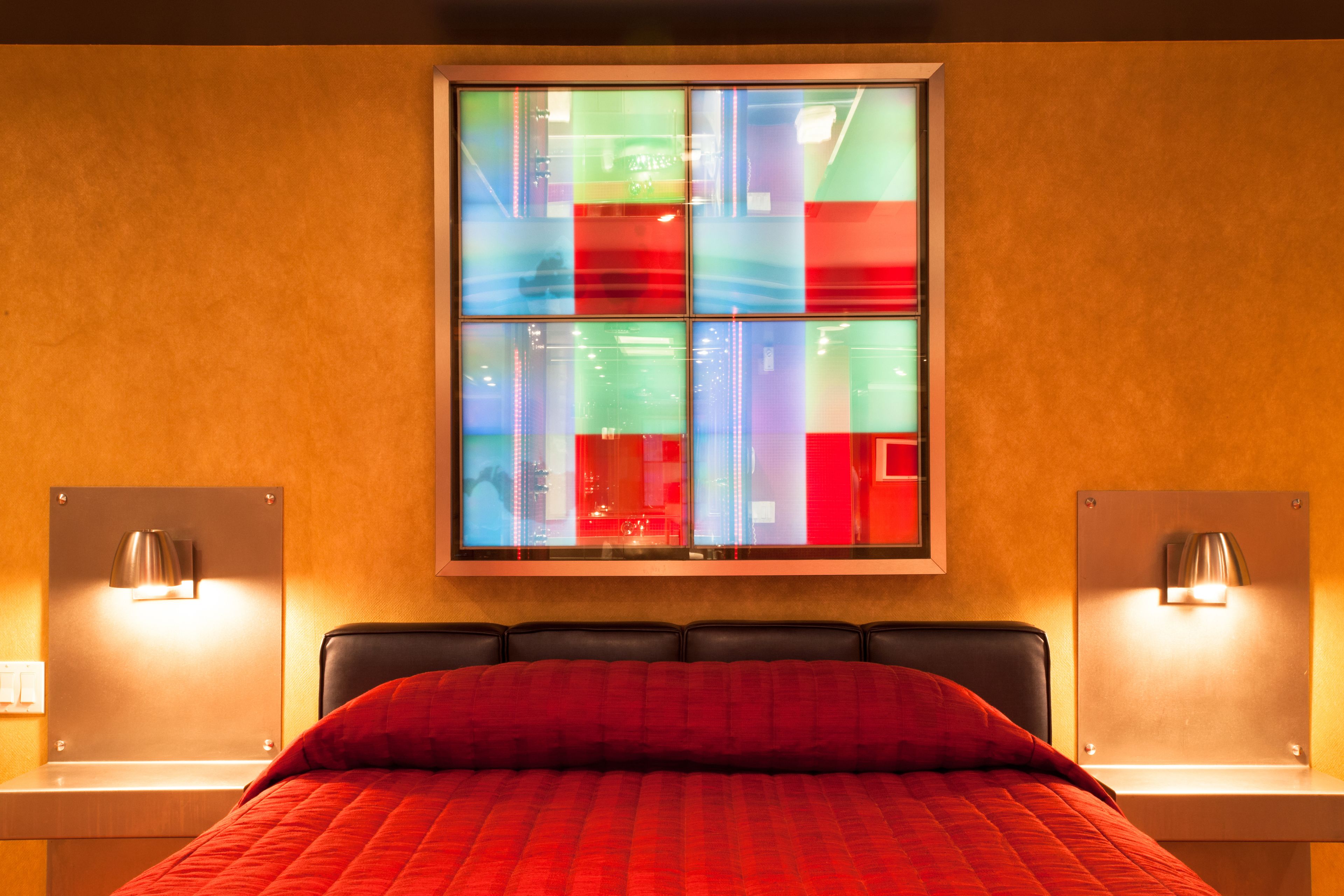 A hotel room with a large bed, two bedside tables, a window with colorful glass panes, and wall-mounted lamps.