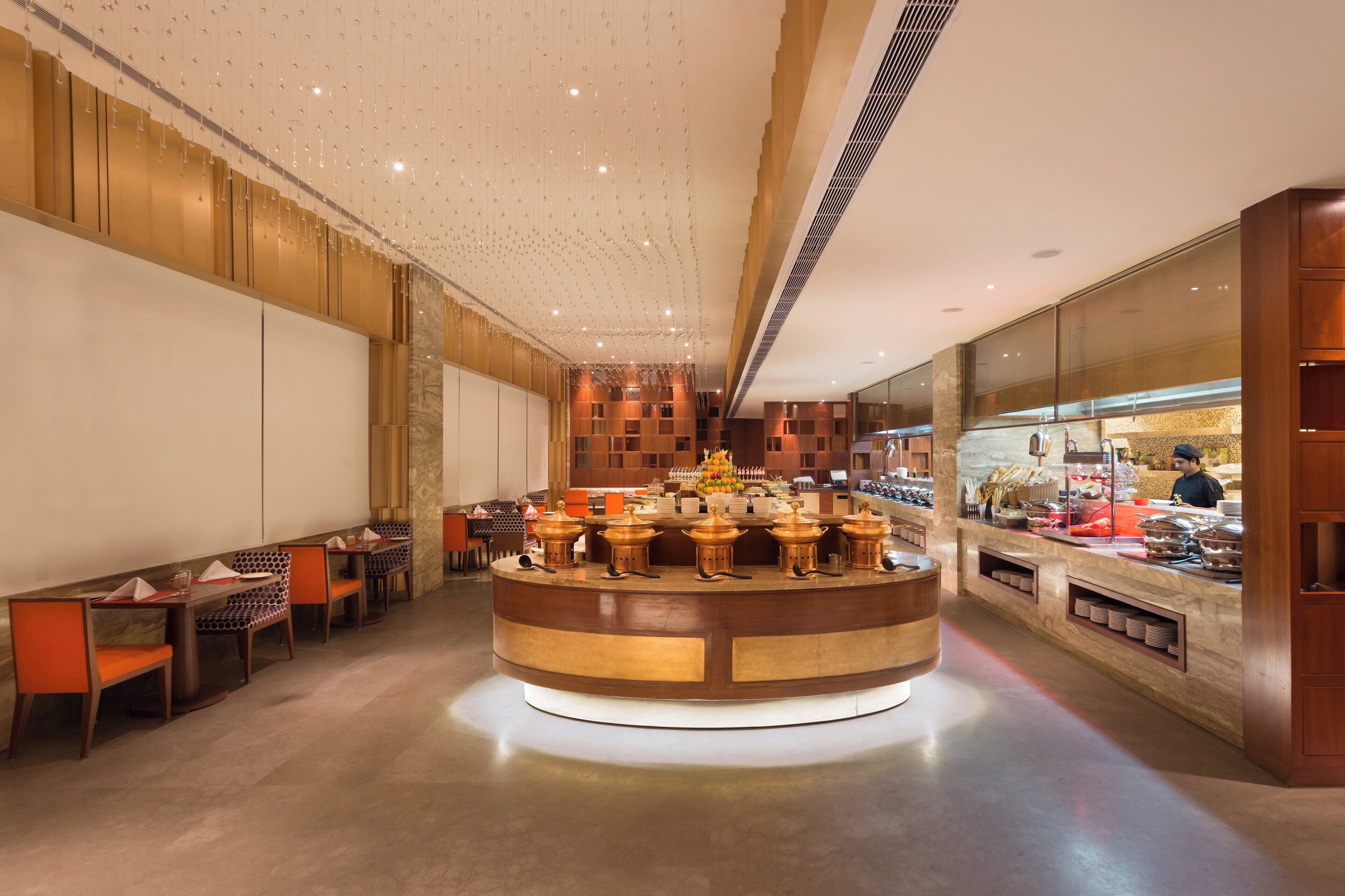 A modern restaurant interior with a central buffet area, seating arrangements, and a kitchen visible through a glass partition.