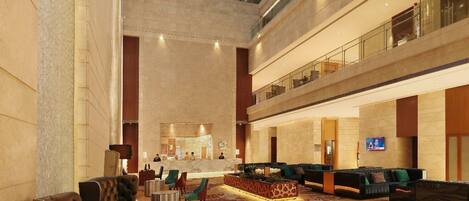 A spacious lobby with a high ceiling, a central reception area, multiple seating arrangements, and a glass railing on the upper level.