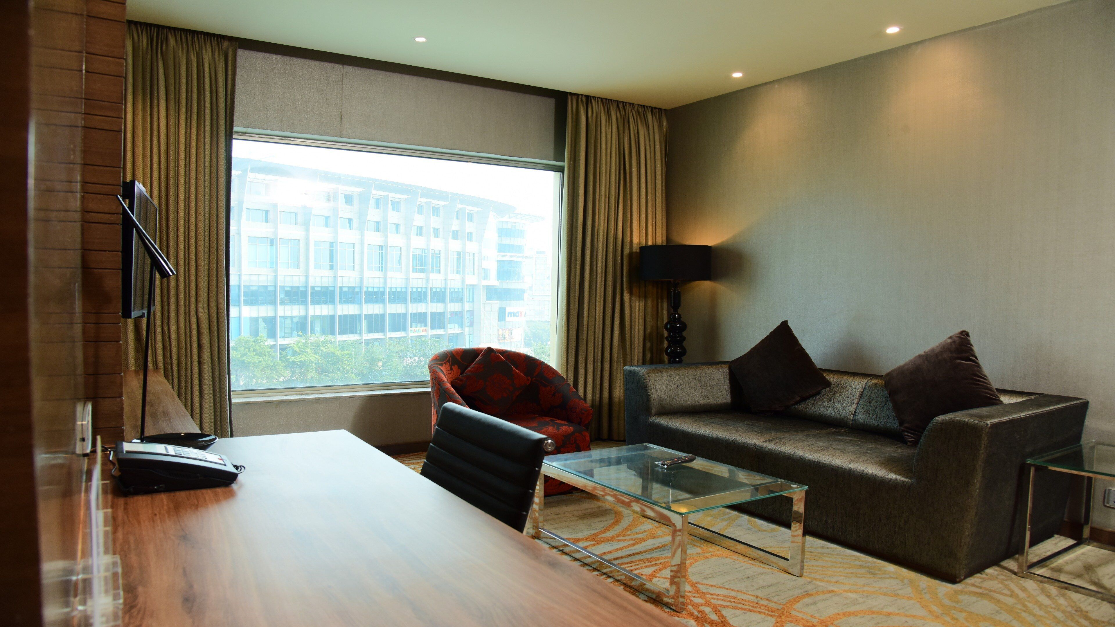 A hotel room with a large window, a sofa, a glass coffee table, a desk, and a lamp.