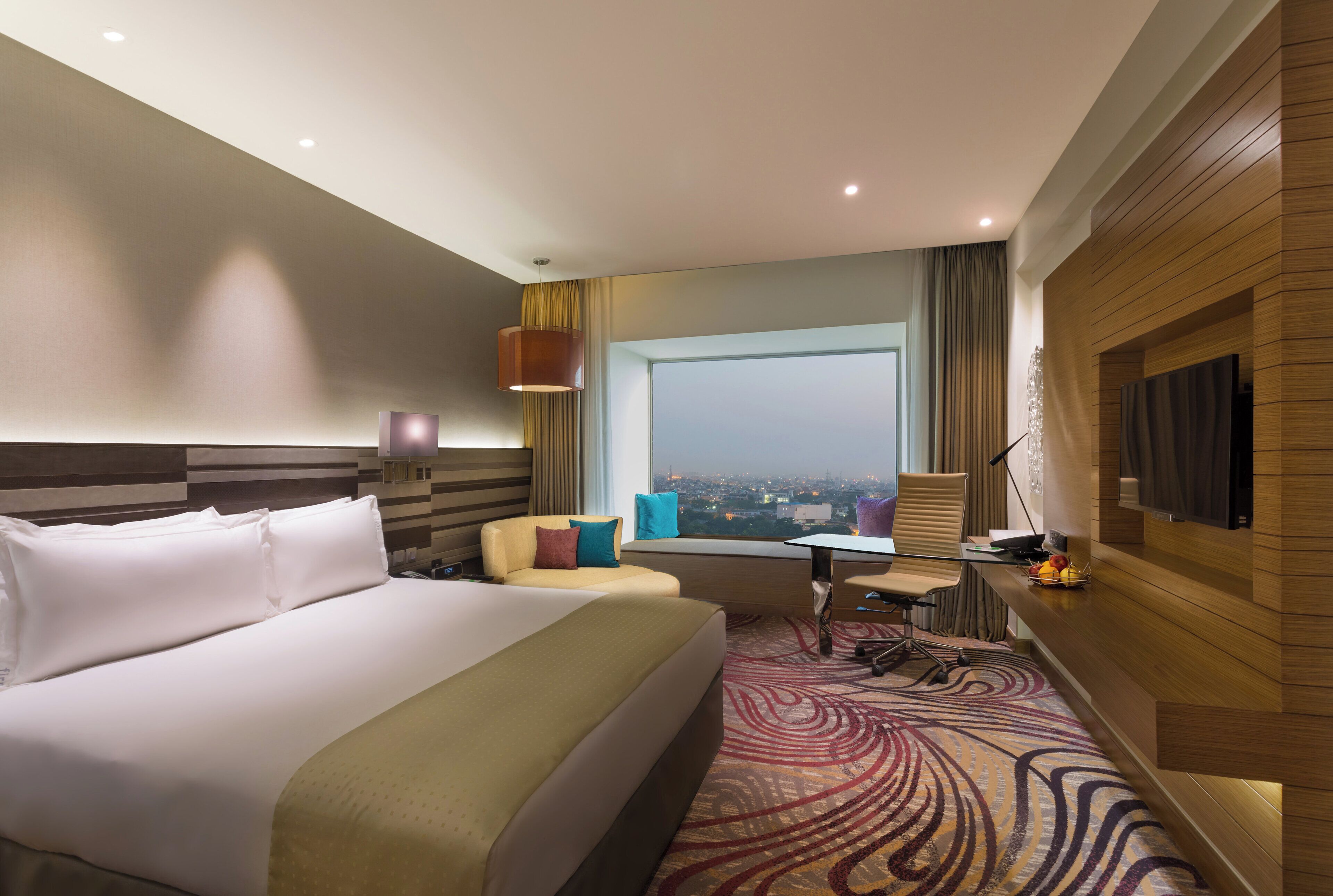 A hotel room with a large bed, a desk, a chair, a small table, a TV, and a view of the city.