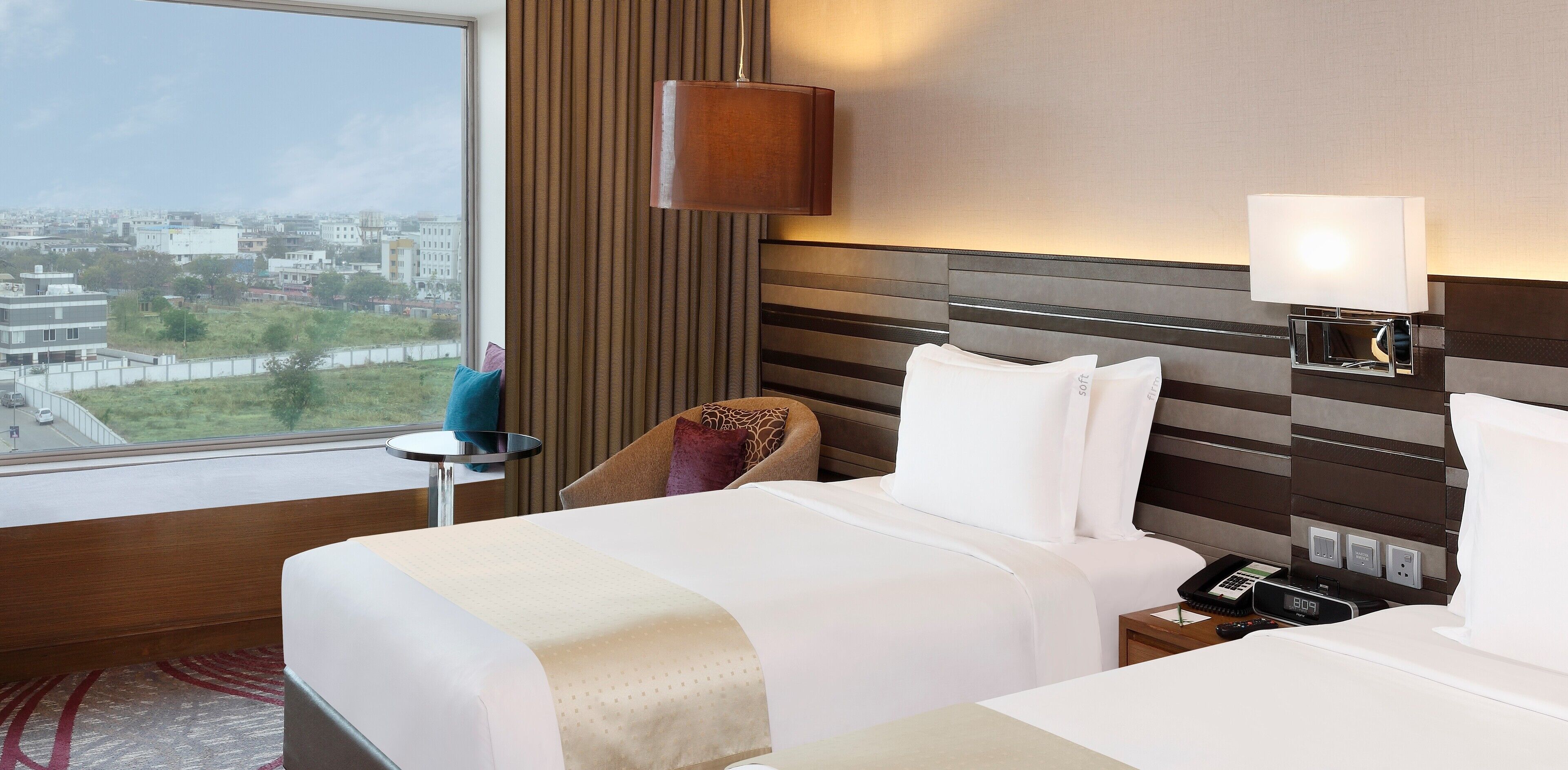 A hotel room with a large window, a bed, a chair, a bedside table, and a lamp.