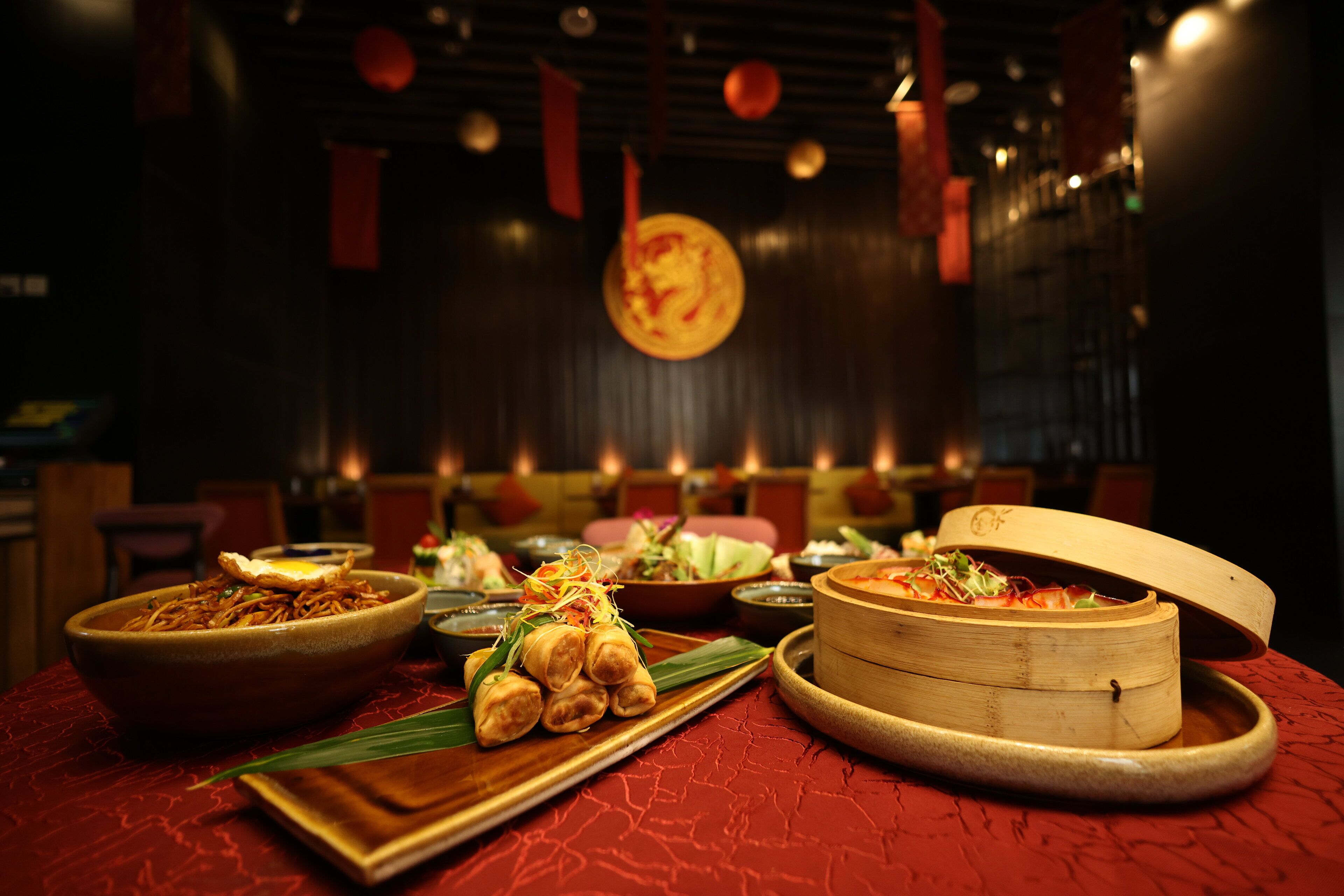 A dining table with various dishes, including a bowl of noodles, a plate of spring rolls, and a bamboo steamer.