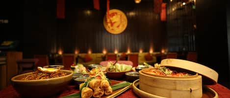 A dining table with various dishes, including a bowl of noodles, a plate of spring rolls, and a bamboo steamer.