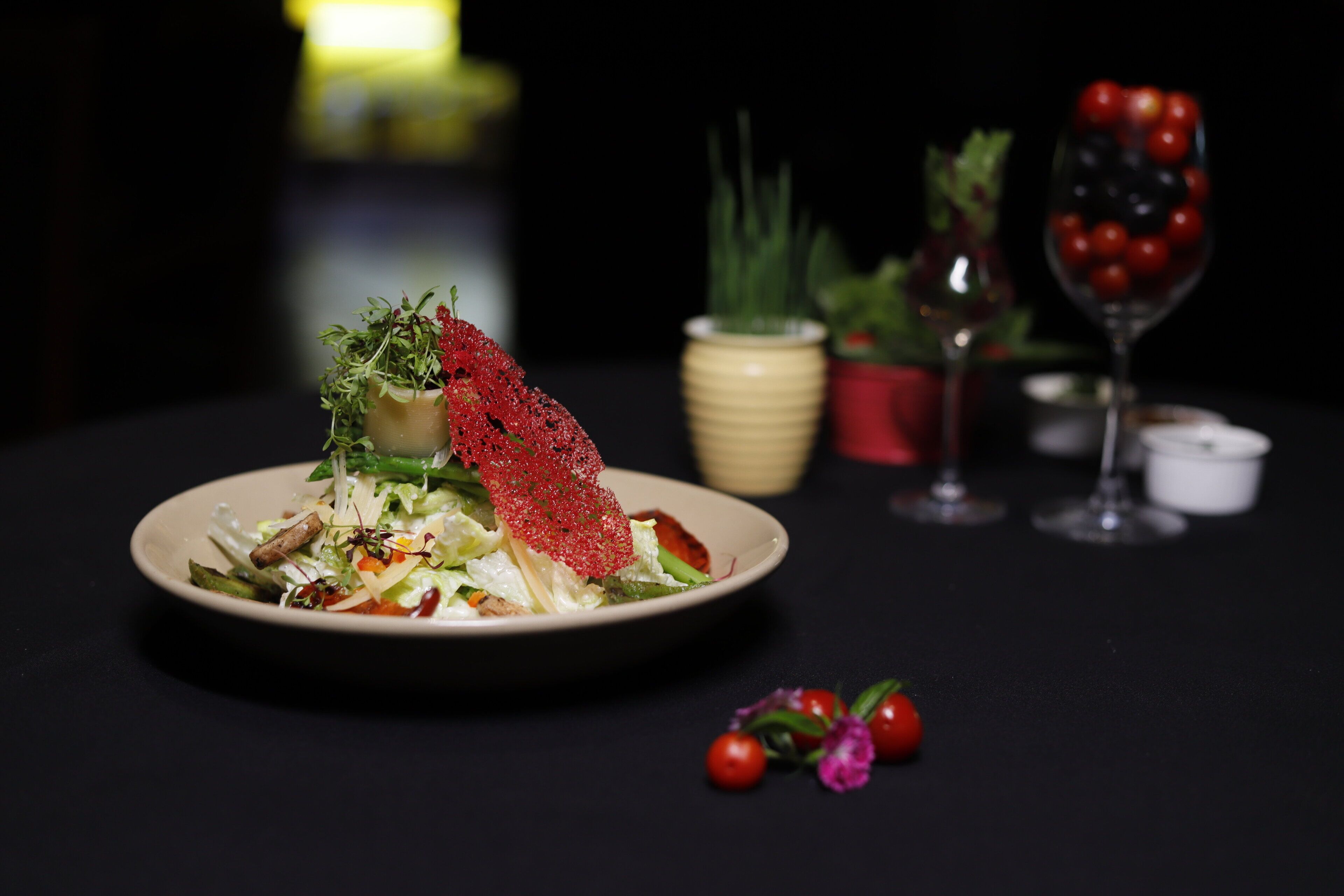 A plate of food with a decorative red element, a glass of red wine, and a small vase with green plants.