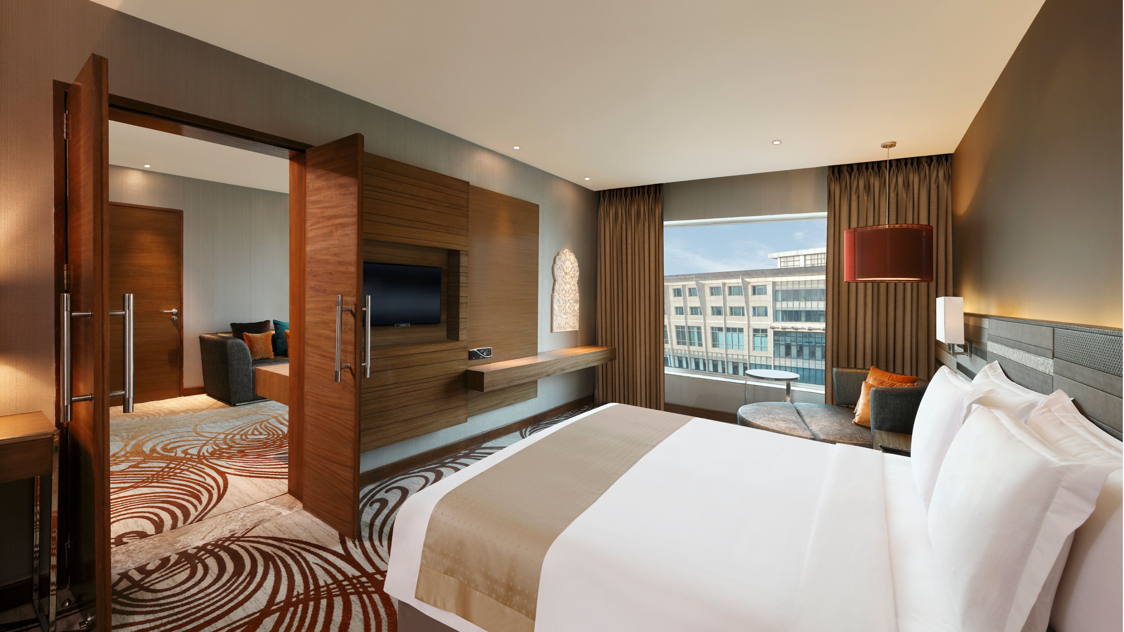 A modern hotel room with a large bed, a TV mounted on the wall, a seating area with a table, and a view of the cityscape through the window.