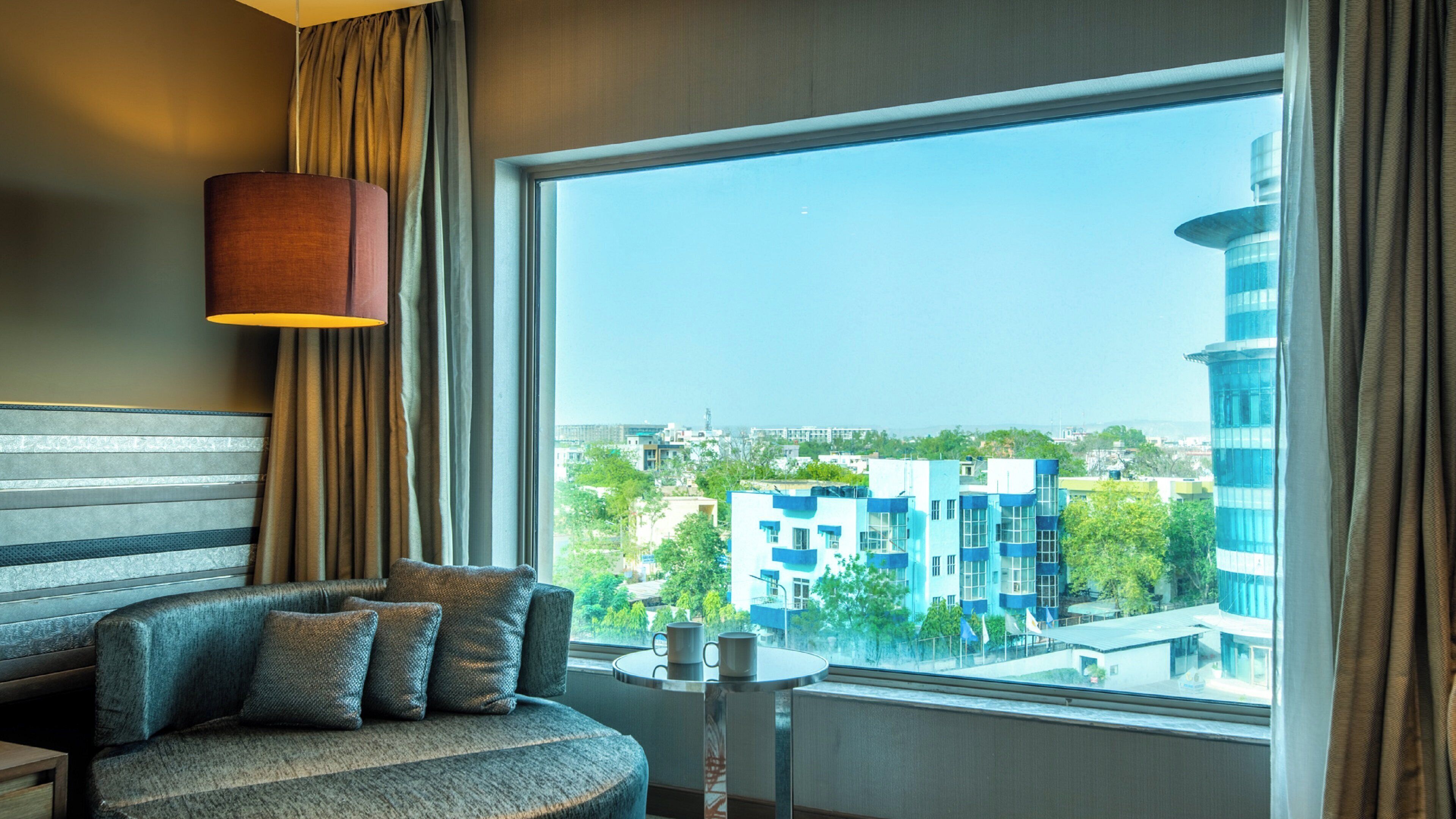 A hotel room with a large window, a sofa, a small table, and a lamp.