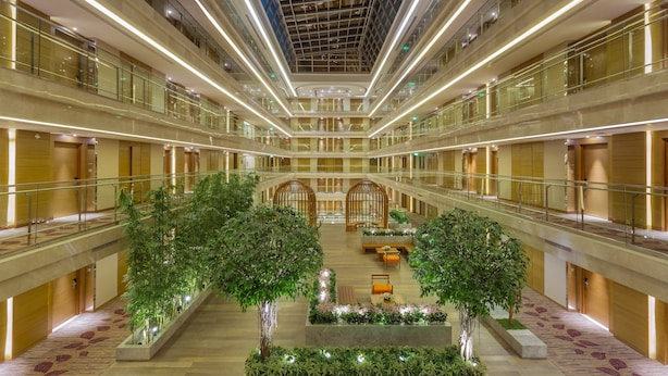 Un atrium à plusieurs niveaux doté de rampes en verre, un jardin central agrémenté d’arbres et un design de plafond moderne.