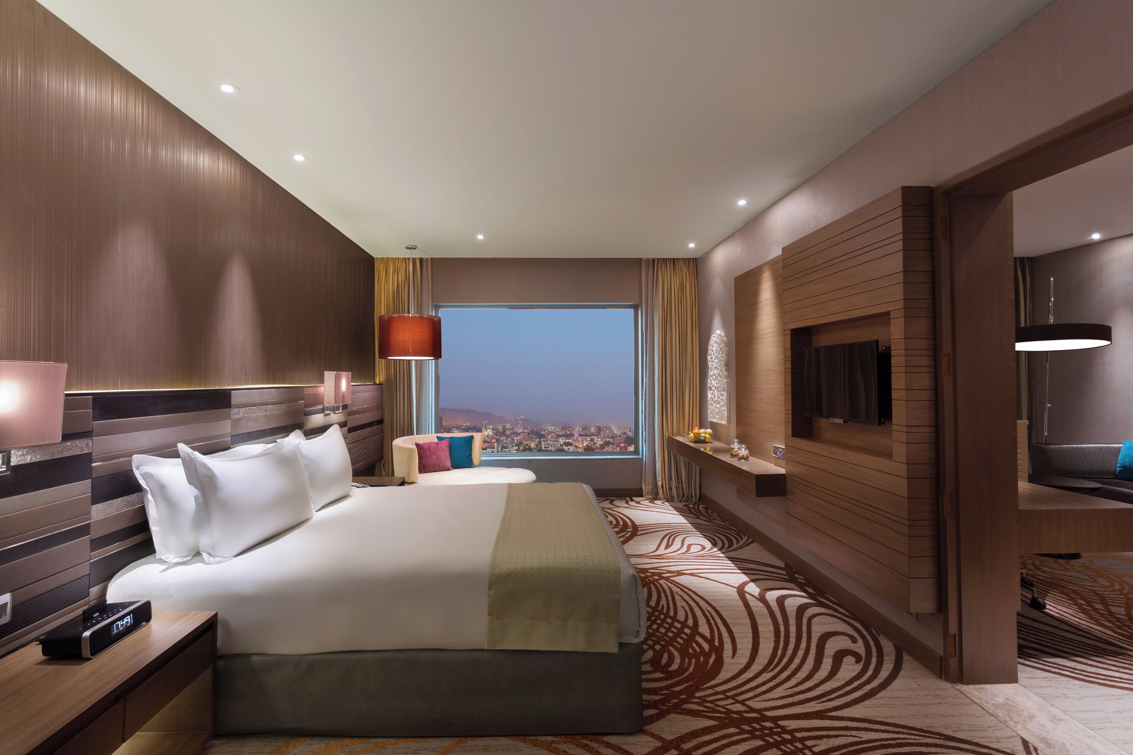A hotel room with a large bed, a bedside table, a lamp, a TV, and a view of a cityscape.
