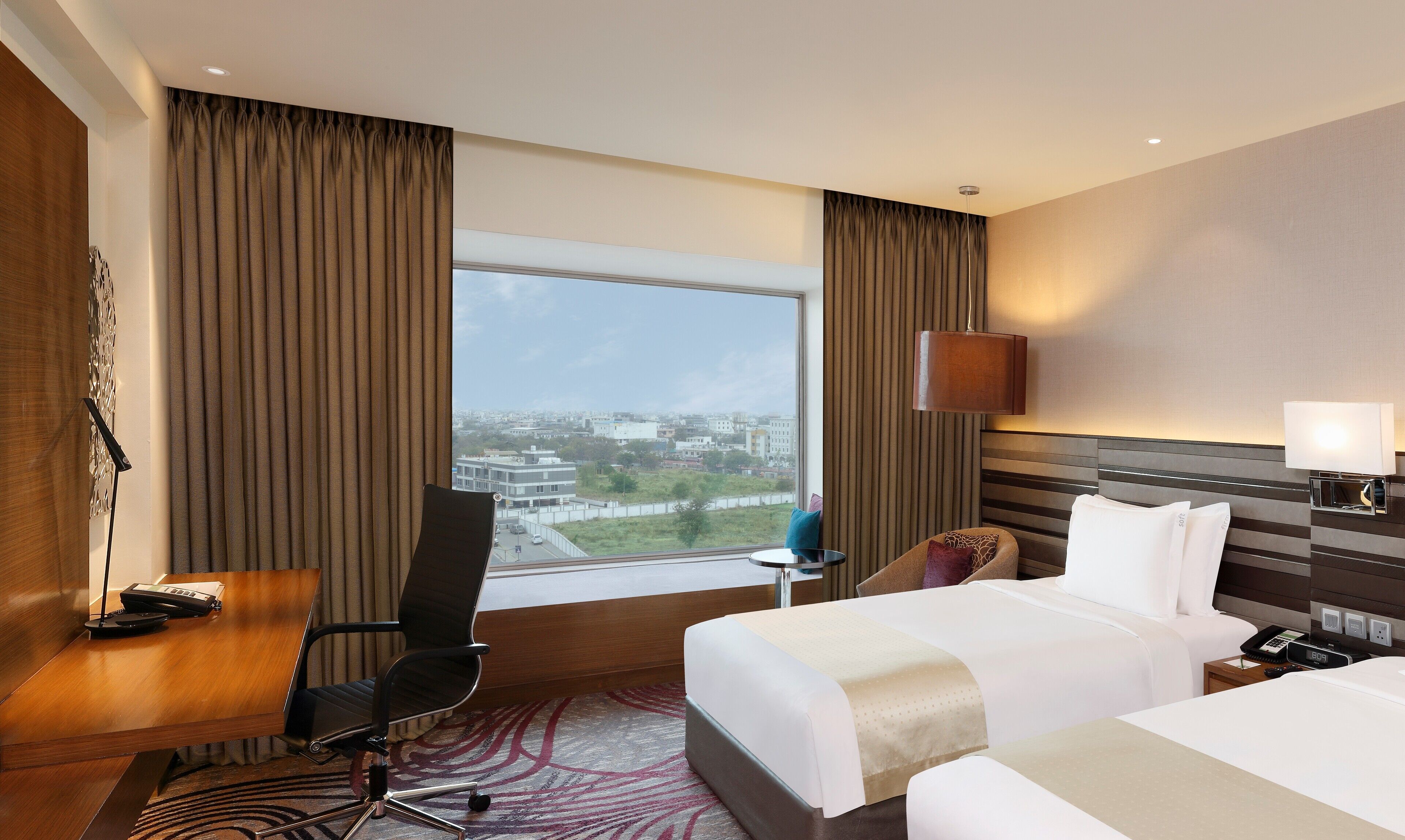 A hotel room with a desk, two beds, a view of the city, and a modern lamp.