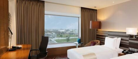 Habitación de hotel con escritorio, dos camas, vistas a la ciudad y una lámpara moderna.