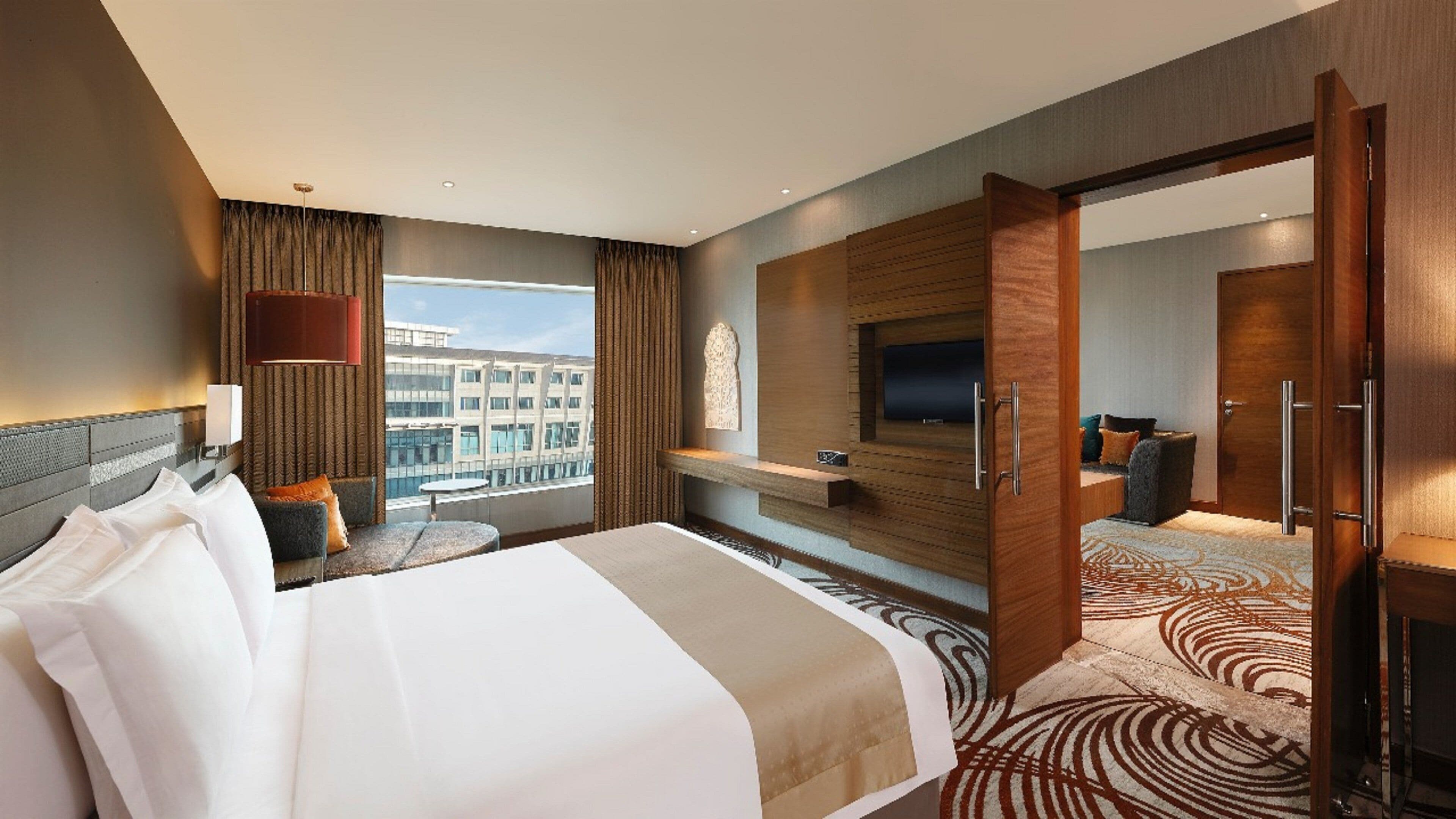 A modern hotel room with a large bed, a seating area with a sofa and armchairs, a TV mounted on a wooden panel, and a view of a cityscape through the window.