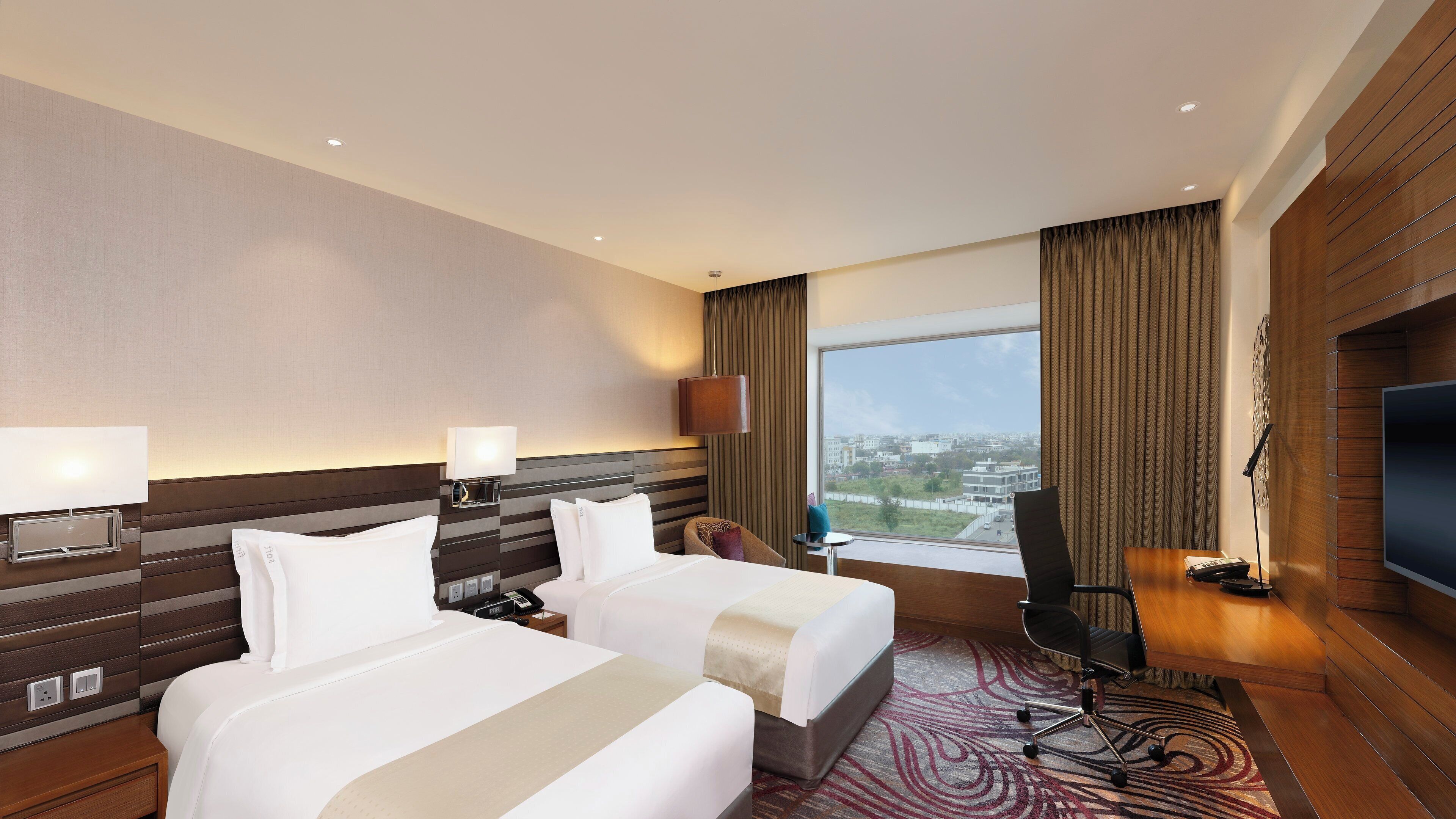 Ein Hotelzimmer mit zwei Betten, einem Fernseher, einem Schreibtisch mit Telefon und Blick auf die Stadt.