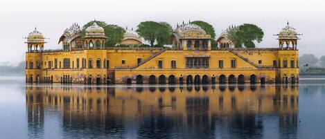 A large, yellow building with multiple domes and arches, reflecting on a calm body of water.