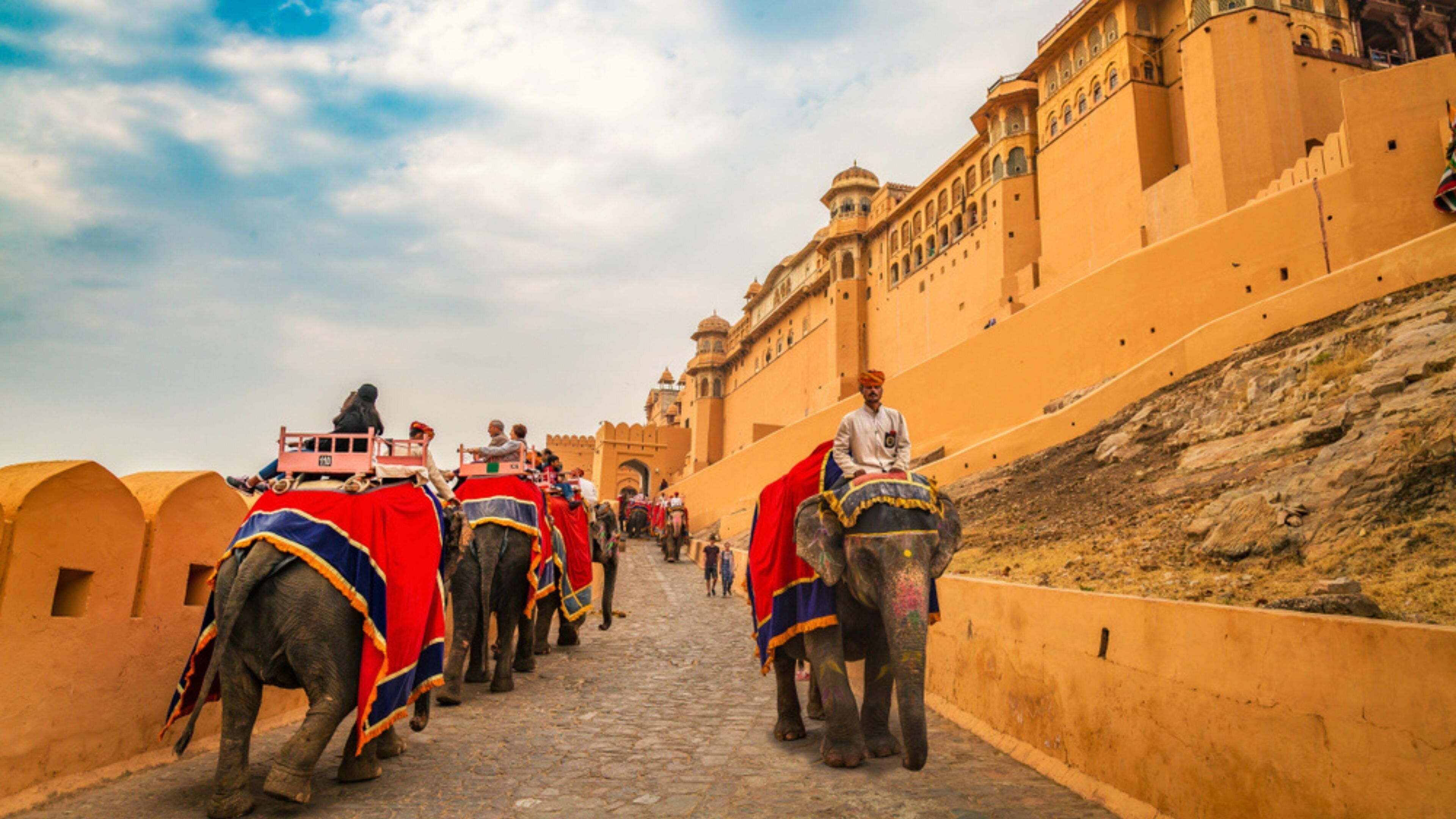 Un groupe d’éléphants, chacun monté par un cavalier, se déplace le long d’un chemin de pierre, leurs dos étant couverts de couvertures colorées. Ils passent à côté d’un grand bâtiment historique, orné d’arches et de coupoles.