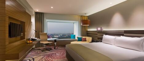 Habitación de hotel con una cama grande, un escritorio con silla, un televisor montado en la pared y vistas a la ciudad desde la ventana.