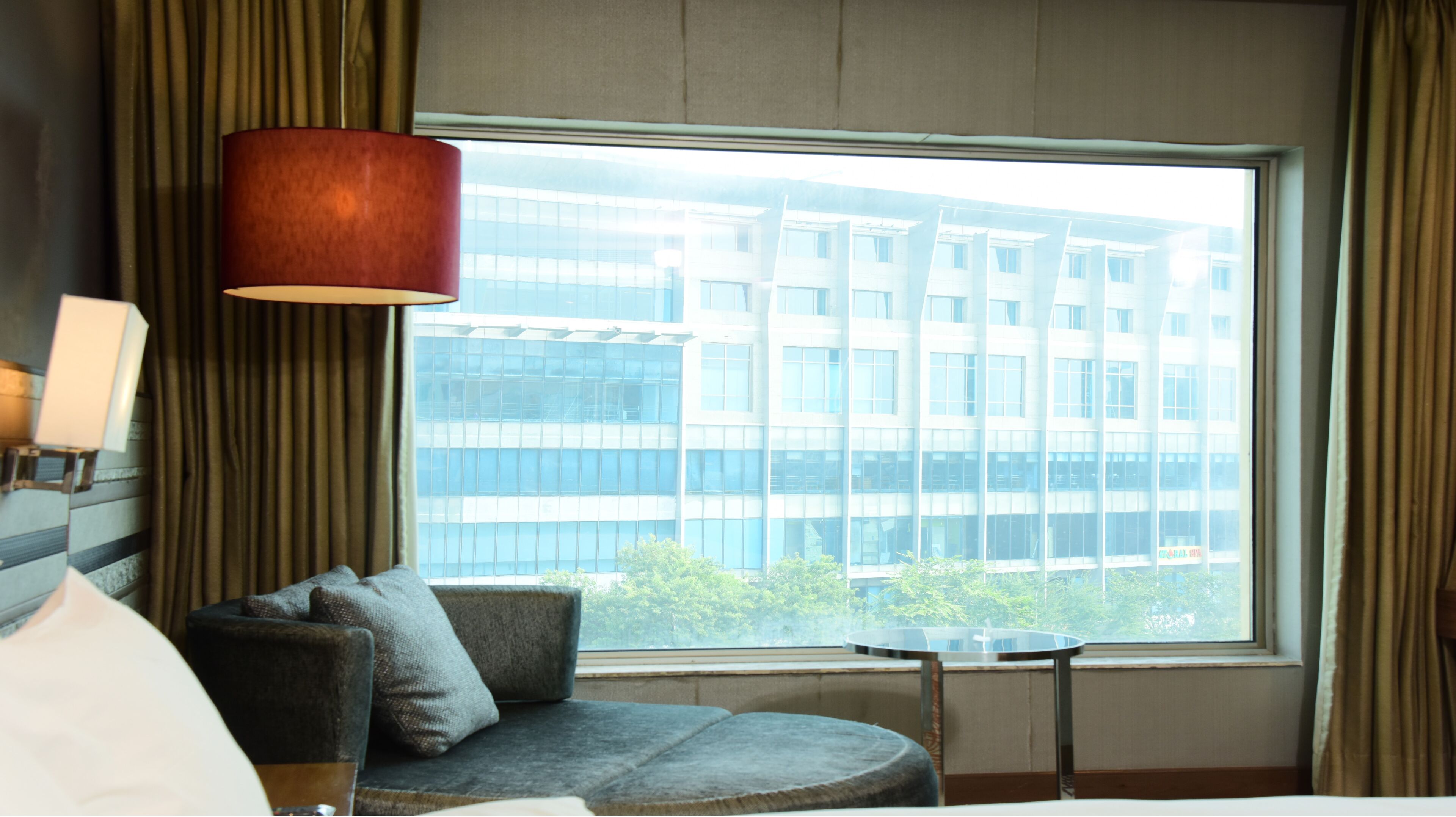 A hotel room with a large window, a sofa, a small table, and a lamp.