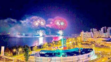 Une vue sur le front de mer illuminée par des feux d’artifice, un grand bâtiment circulaire et un paysage urbain en arrière-plan.