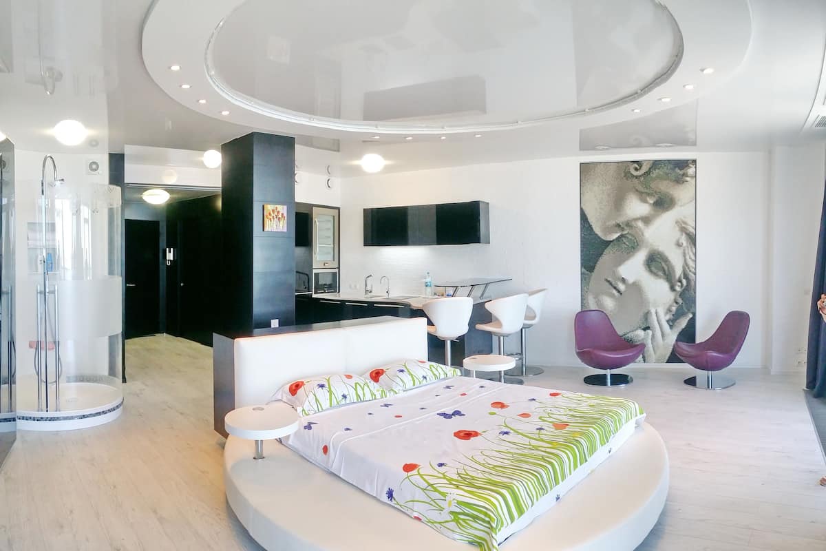 A modern hotel room with a large bed, a seating area with two chairs, a TV mounted on the wall, and a bathroom with a glass shower enclosure.