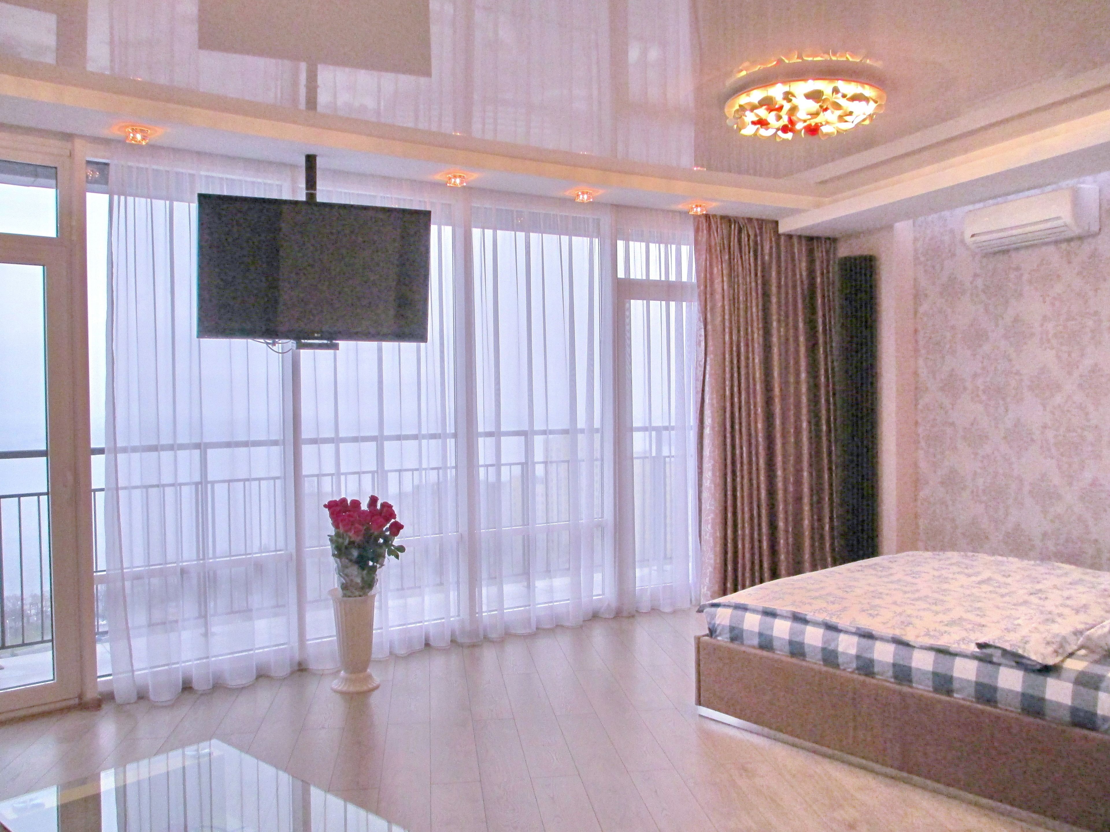 Habitación de hotel con una cama grande, un televisor de pantalla plana en la pared, un jarrón con flores y ventanas amplias con cortinas traslúcidas.