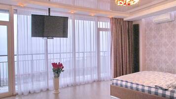 Habitación de hotel con una cama grande, un televisor de pantalla plana en la pared, un jarrón con flores y ventanas amplias con cortinas traslúcidas.