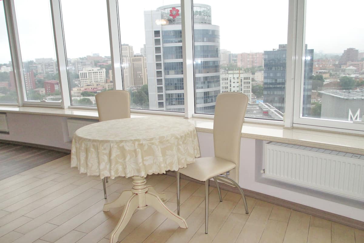 A room with a round table and two chairs, large windows with a city view, and a radiator on the wall.