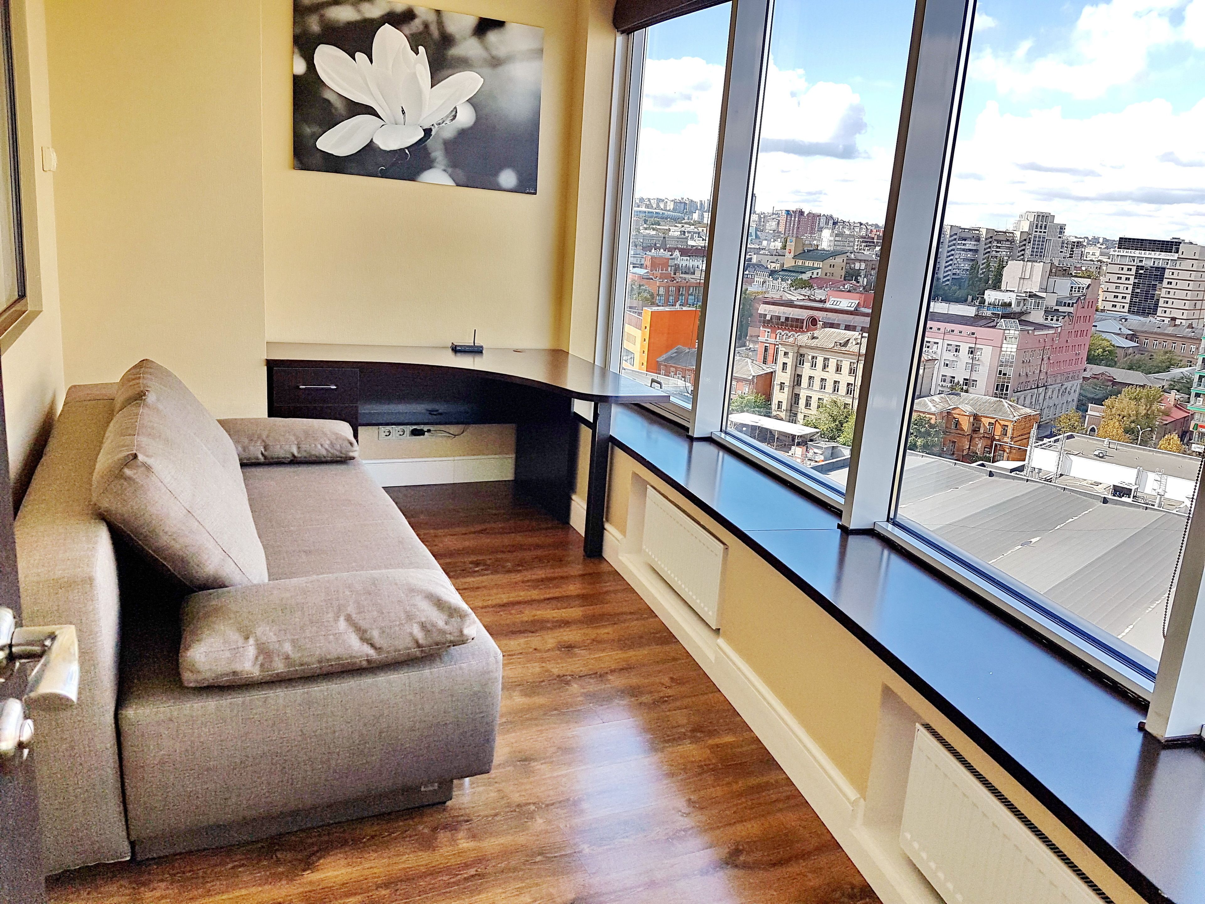 Ein modernes Wohnzimmer mit einem grauen Sofa, einem großen Fenster mit Blick auf die Stadt und einem an der Wand hängenden Bild von einer Blume.