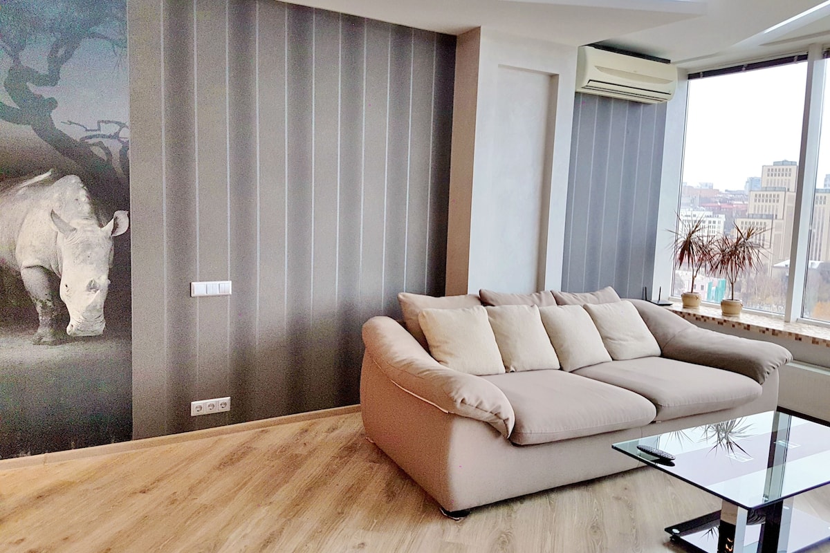 A modern living room with a beige sofa, a glass coffee table, and a city view through large windows.